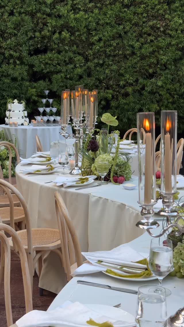 A long wedding table decorated with greenery centerpieces and tall lit candles against a hedge.