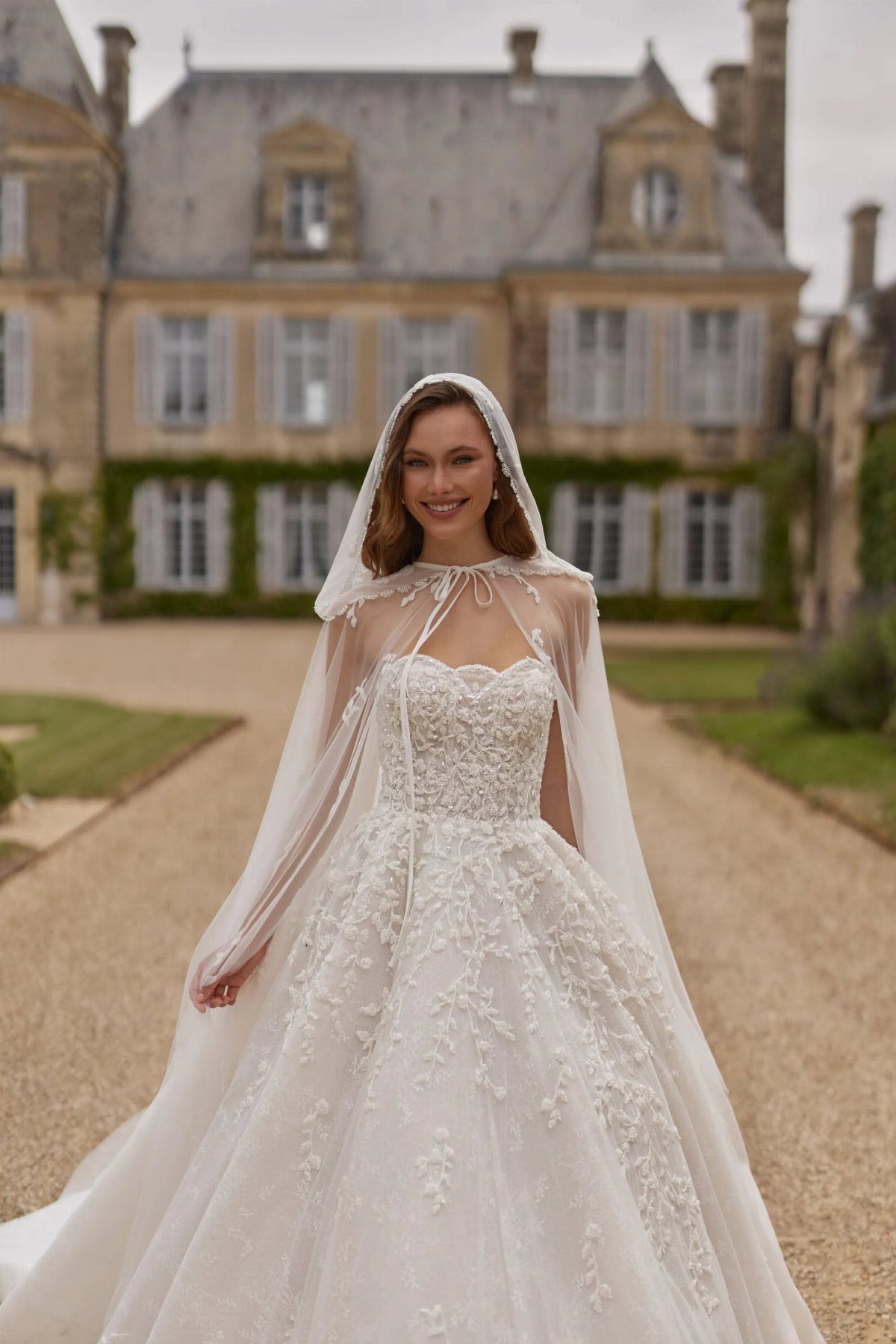 Hooded Wedding Dress: 6 Ideas from Vintage to Modern 5 A woman in a lace wedding dress wearing a sheer white hooded cape outdoors.