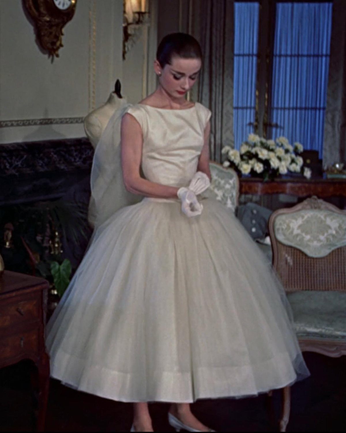 Iconic Wedding Dresses and 6 Stories They Still Tell 2 A woman in a white sleeveless tea-length gown and gloves standing in an elegant room.