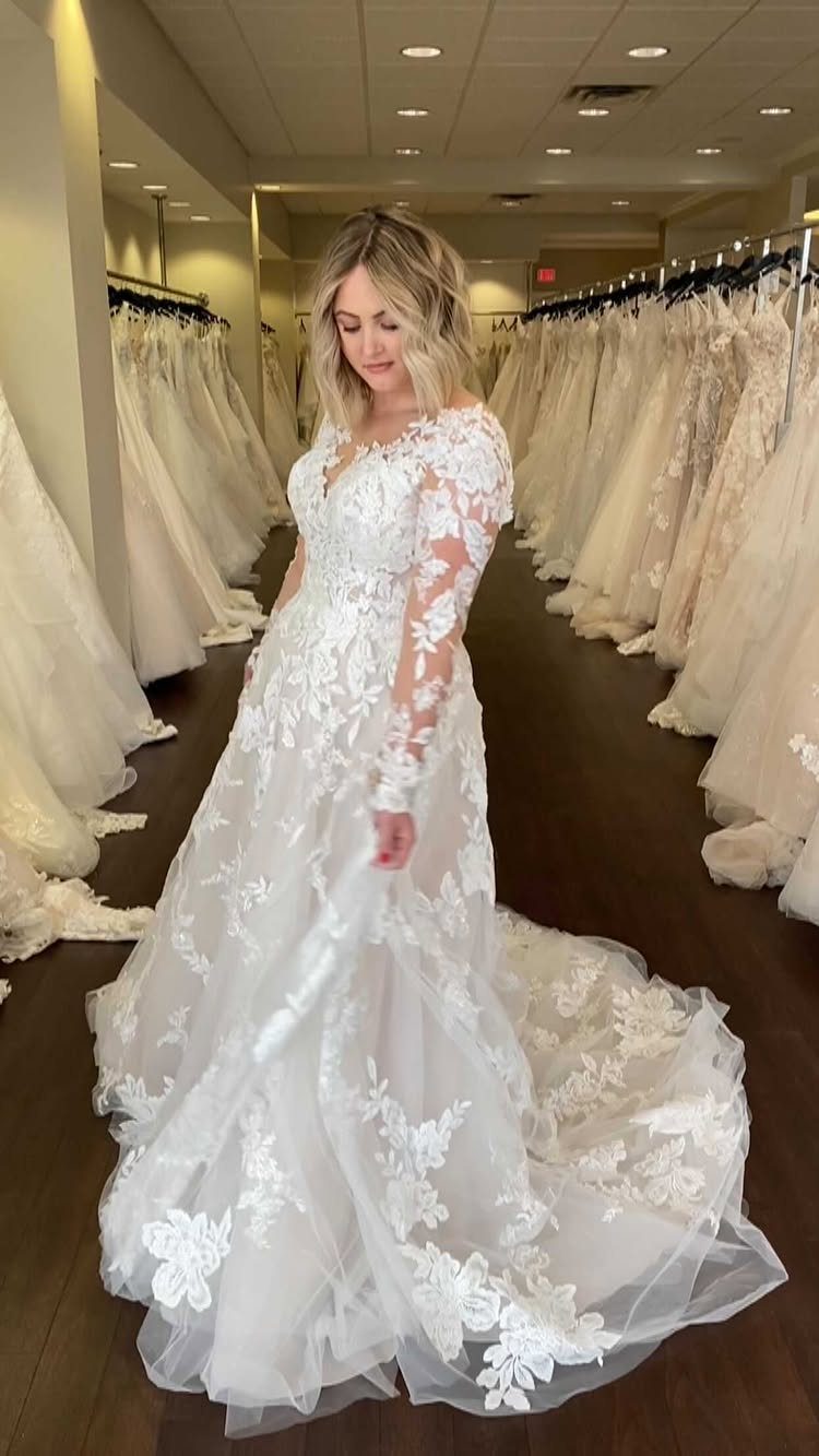 Lace Ball Gowns: 6 Silhouettes and Styles for Your Wedding 2 A woman posing in a white lace ball gown wedding dress with long sleeves in a bridal shop.
