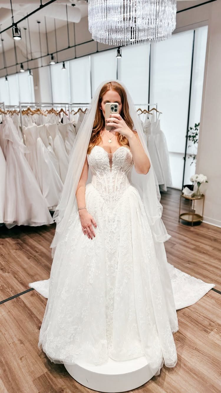 Lace Ball Gowns: 6 Silhouettes and Styles for Your Wedding 5 A woman in a white lace ball gown and veil taking a mirror selfie in a bridal shop.