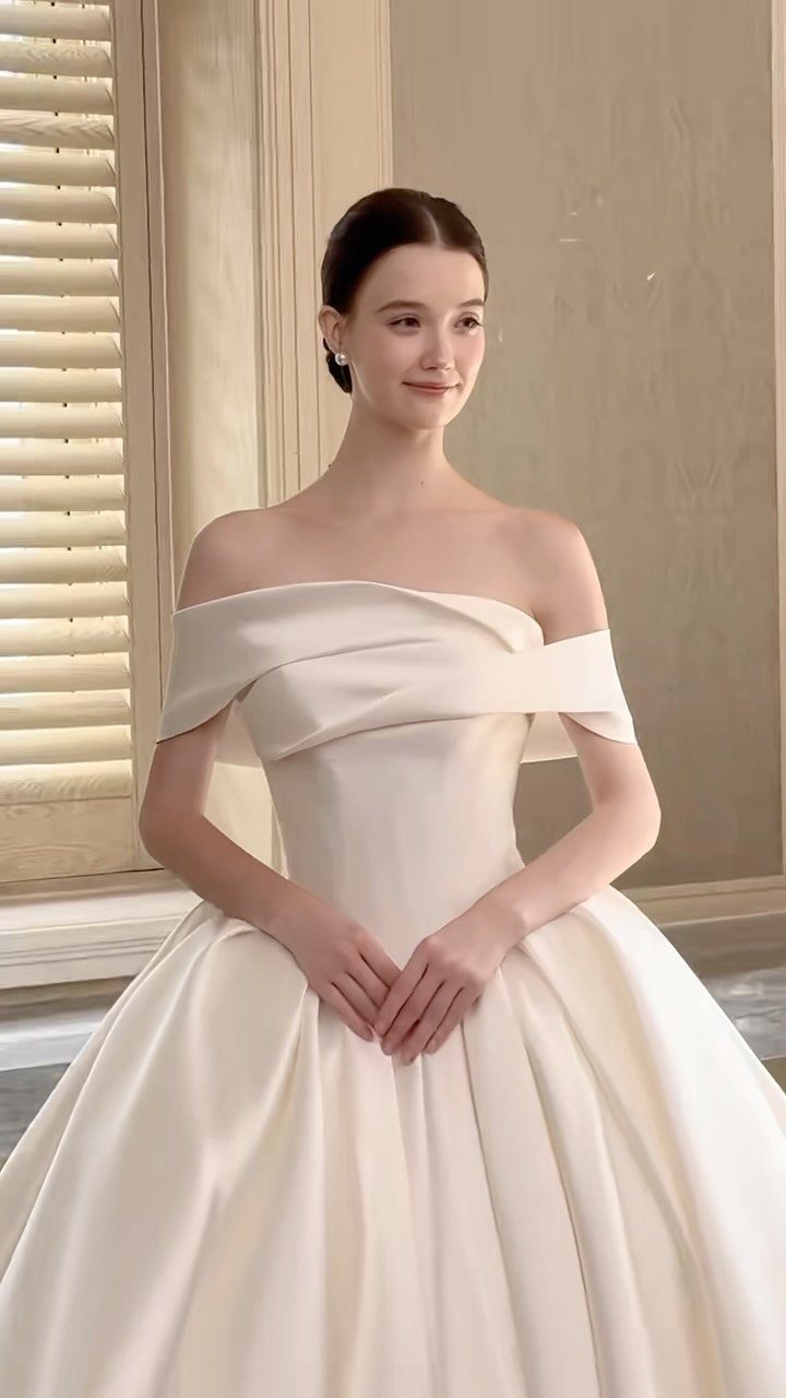 Lace Ball Gowns: 6 Silhouettes and Styles for Your Wedding 4 A woman posing in an elegant off-shoulder ivory satin ball gown wedding dress.