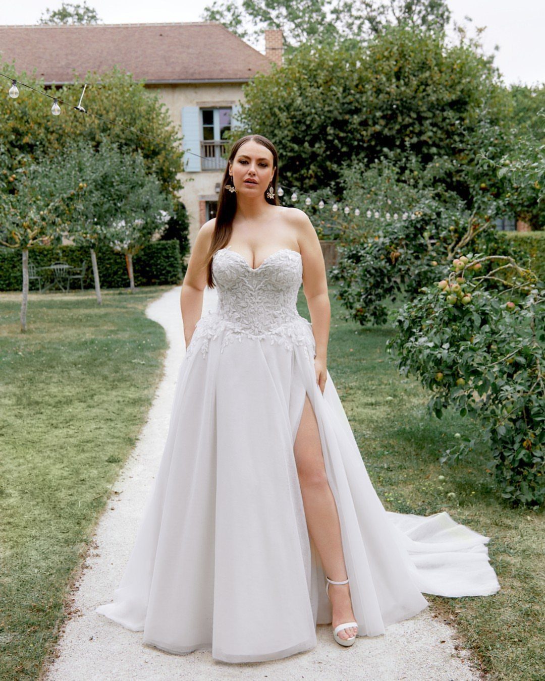 Lace Ball Gowns: 6 Silhouettes and Styles for Your Wedding 6 A woman wearing a white lace strapless ball gown standing on a garden path outdoors.
