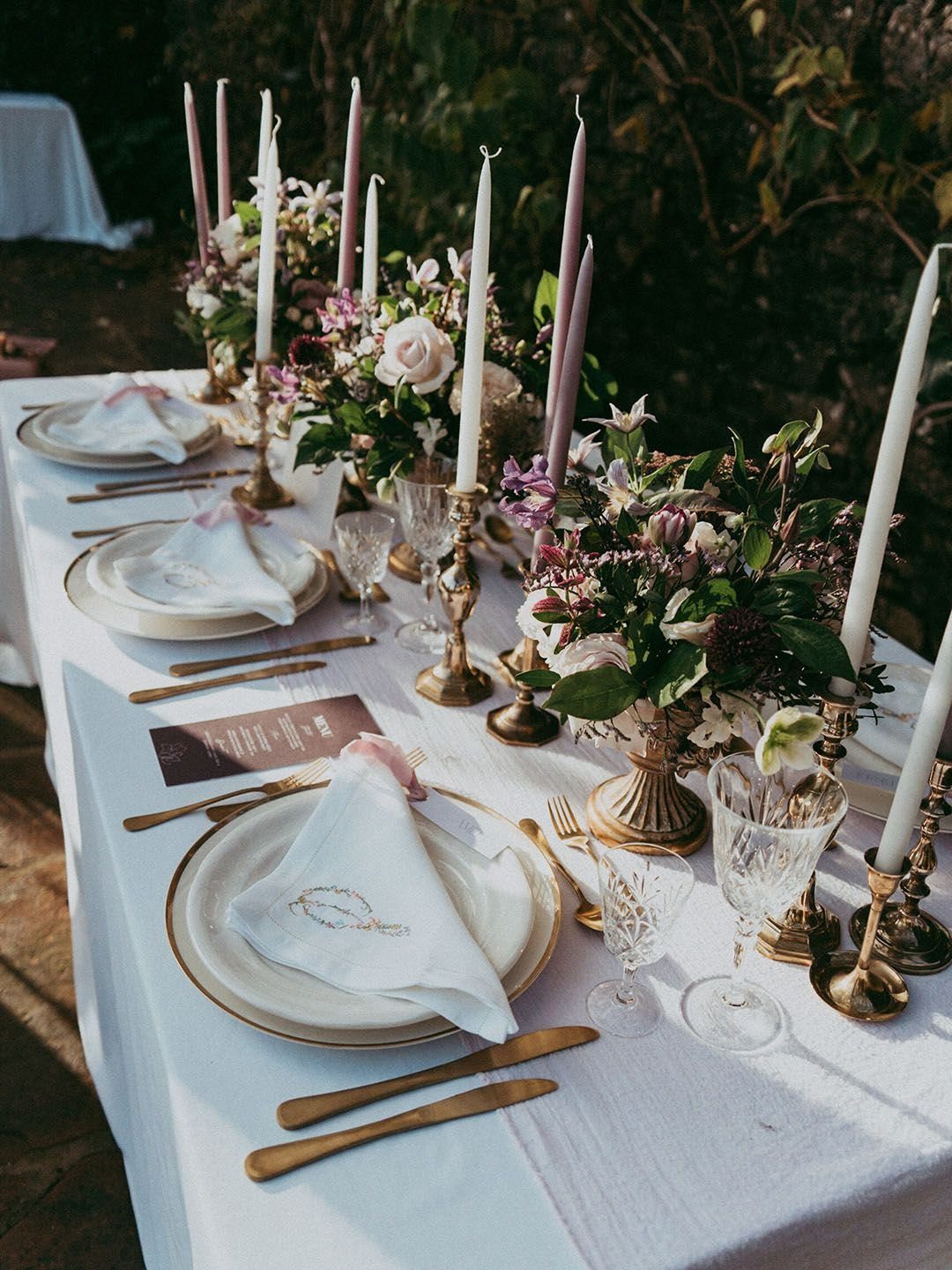 A long wedding table decorated with gold cutlery, white plates, and lavender floral arrangements with tall candles.