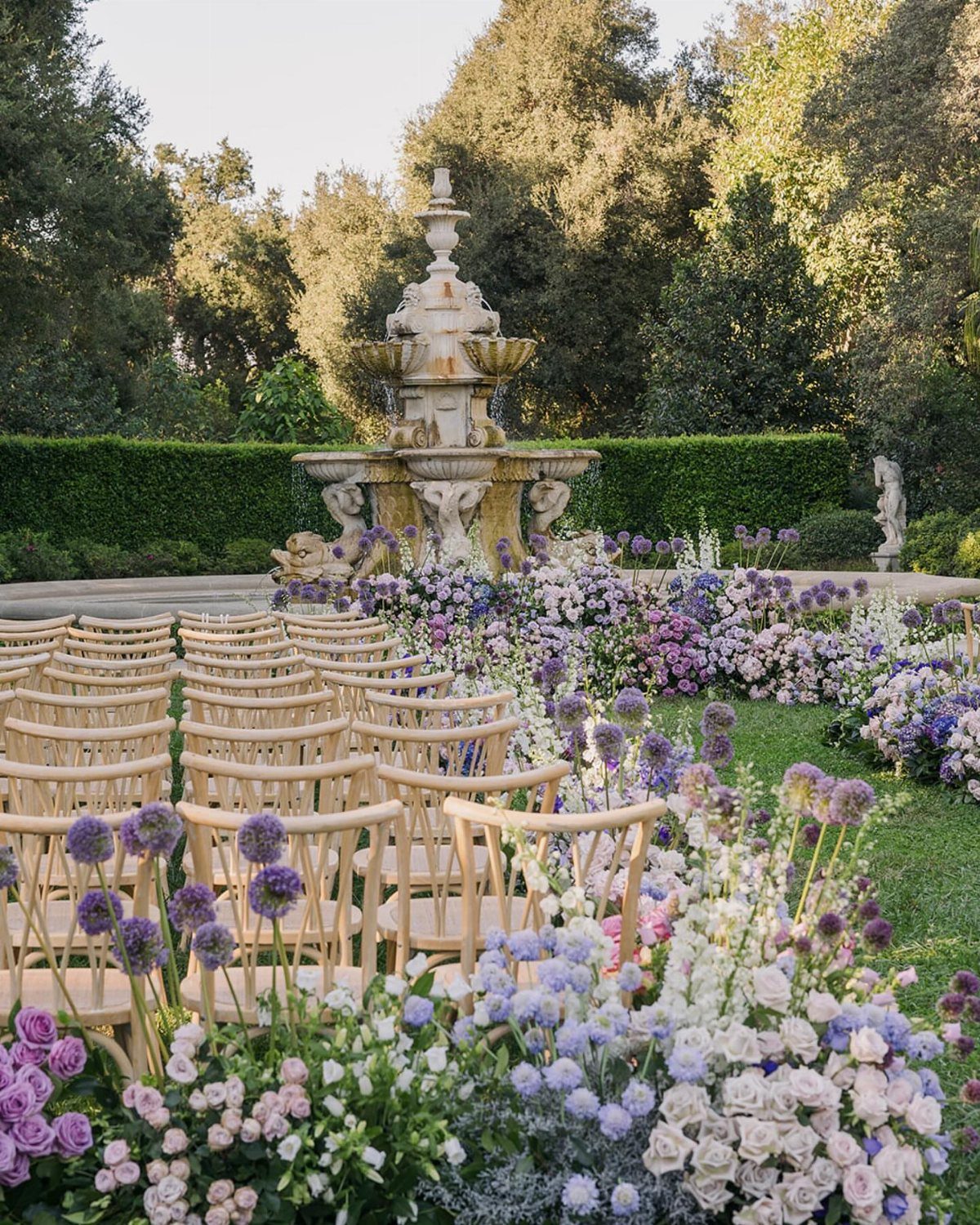 Wooden wedding chairs lined by a lush aisle of purple and white flowers near a fountain.