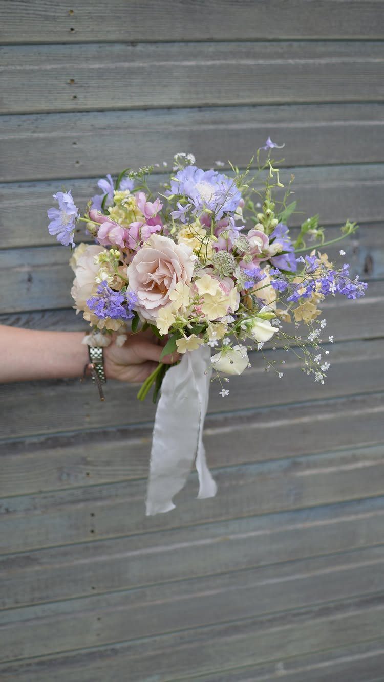 A hand holding a wedding bouquet of lavender and blush pink flowers against wooden planks.