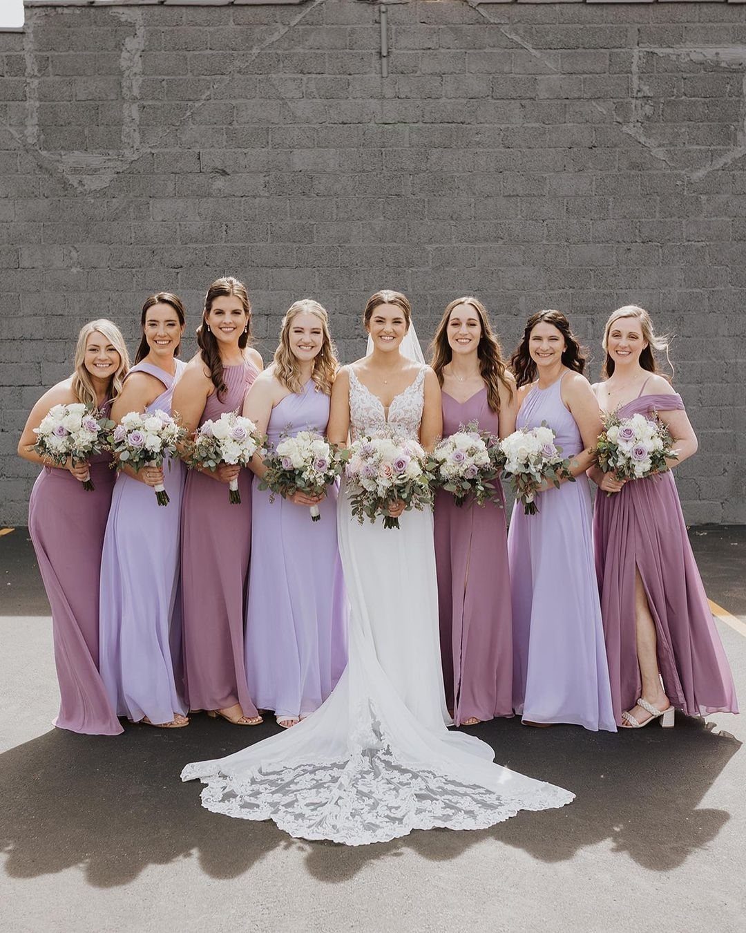 Seven bridesmaids in lavender dresses stand with a bride holding floral bouquets against a grey wall.