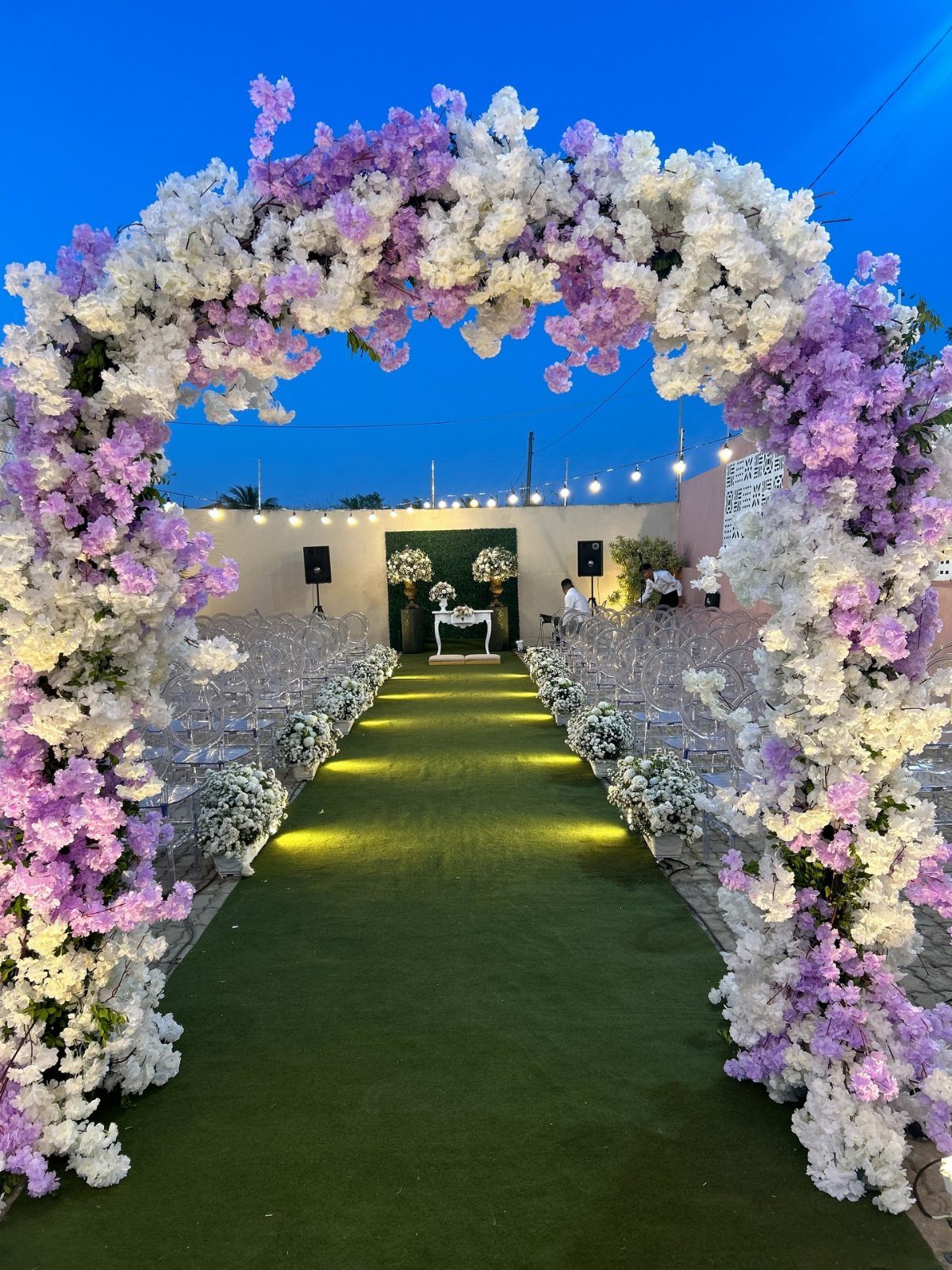 A large arch of white and lavender flowers framing an outdoor wedding ceremony aisle.