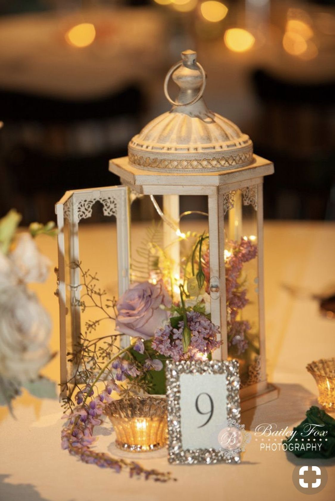 A white decorative lantern filled with glowing lights and purple flowers on a wedding table.