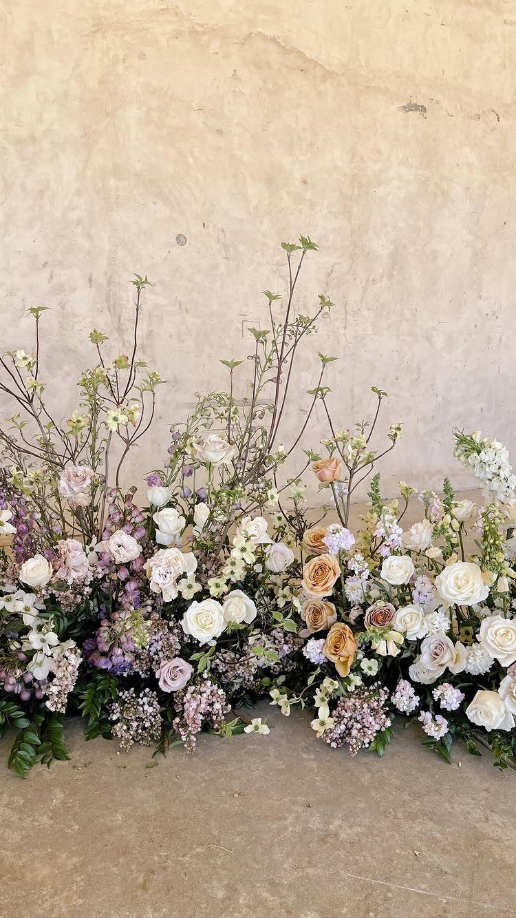Floral arrangement with white, peach, and lavender roses and greenery against a stone wall.