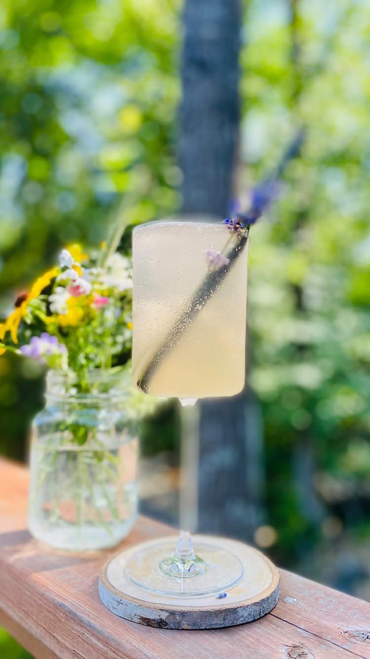 A pale yellow cocktail garnished with a sprig of lavender on a wooden railing.