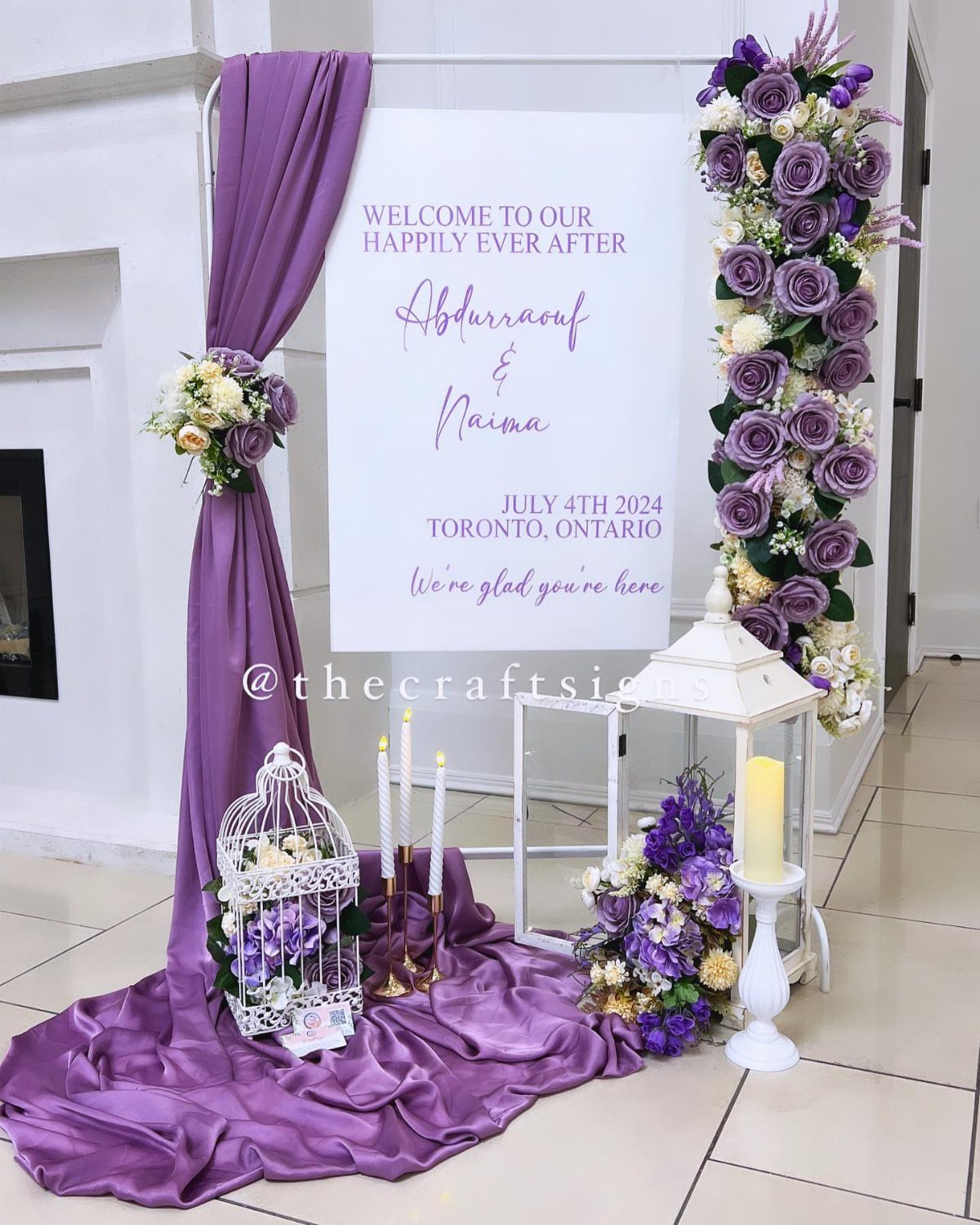 White wedding welcome sign decorated with purple fabric and lavender floral arrangements on a white frame.