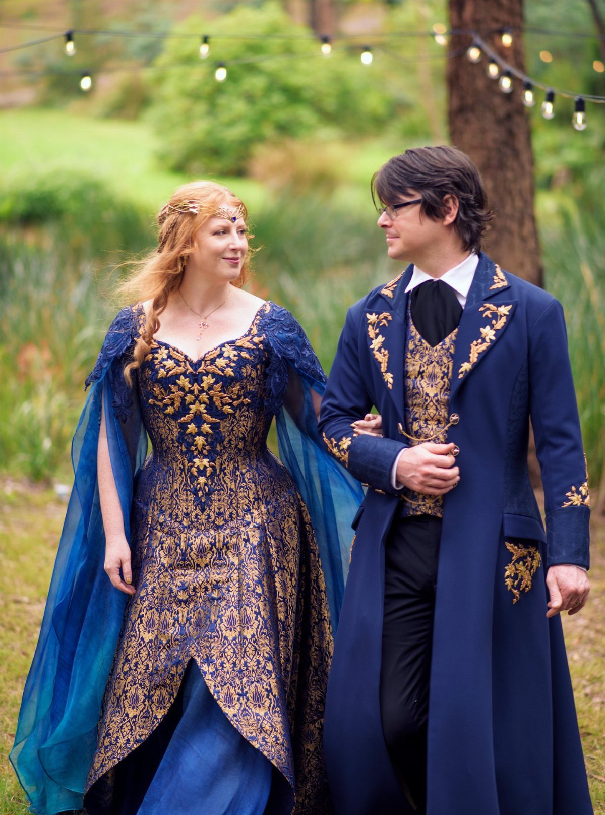 A couple in blue and gold fantasy-style formal attire standing in a wooded outdoor setting.