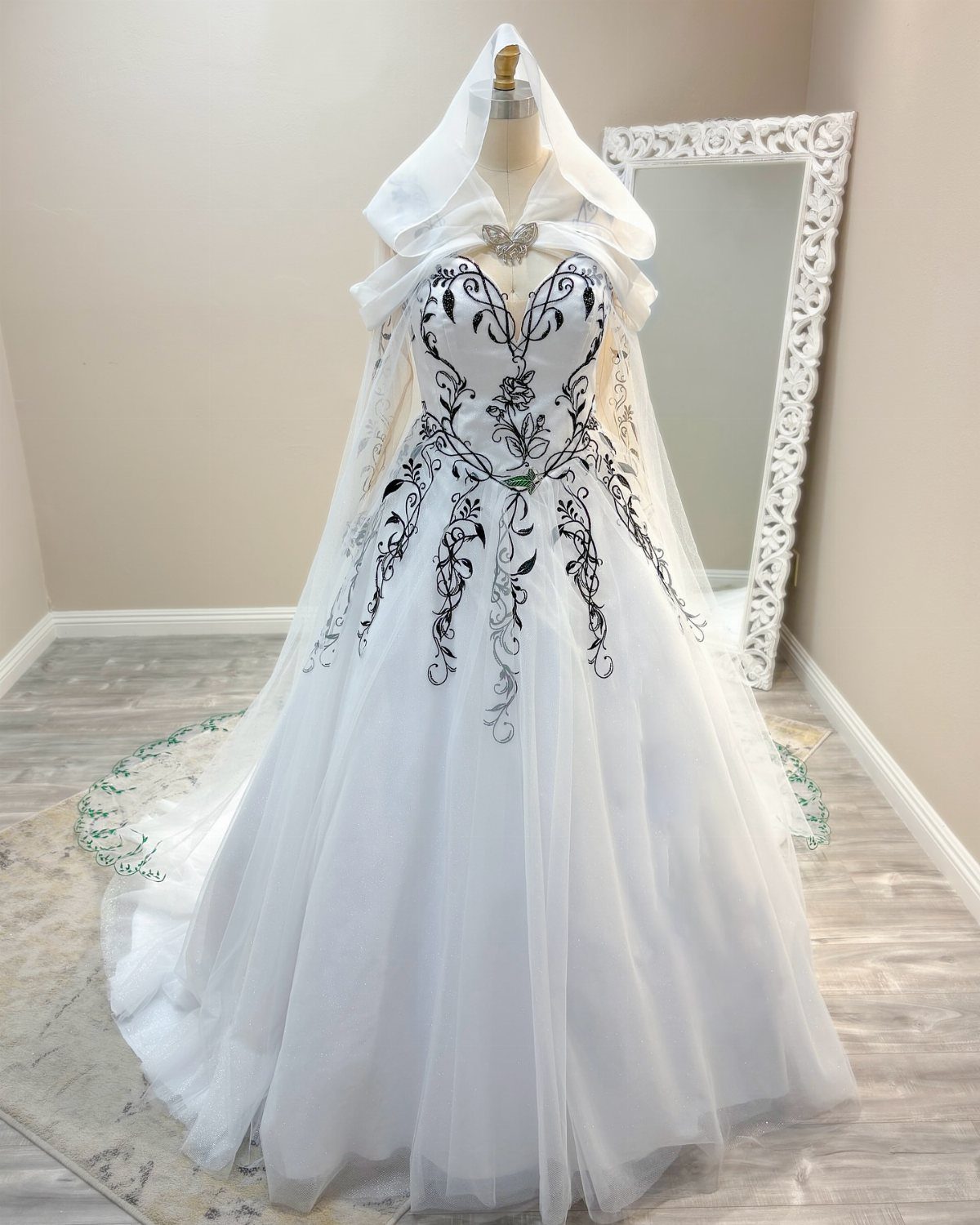 White wedding dress with black vine embroidery and a sheer hooded bridal cape on a mannequin.