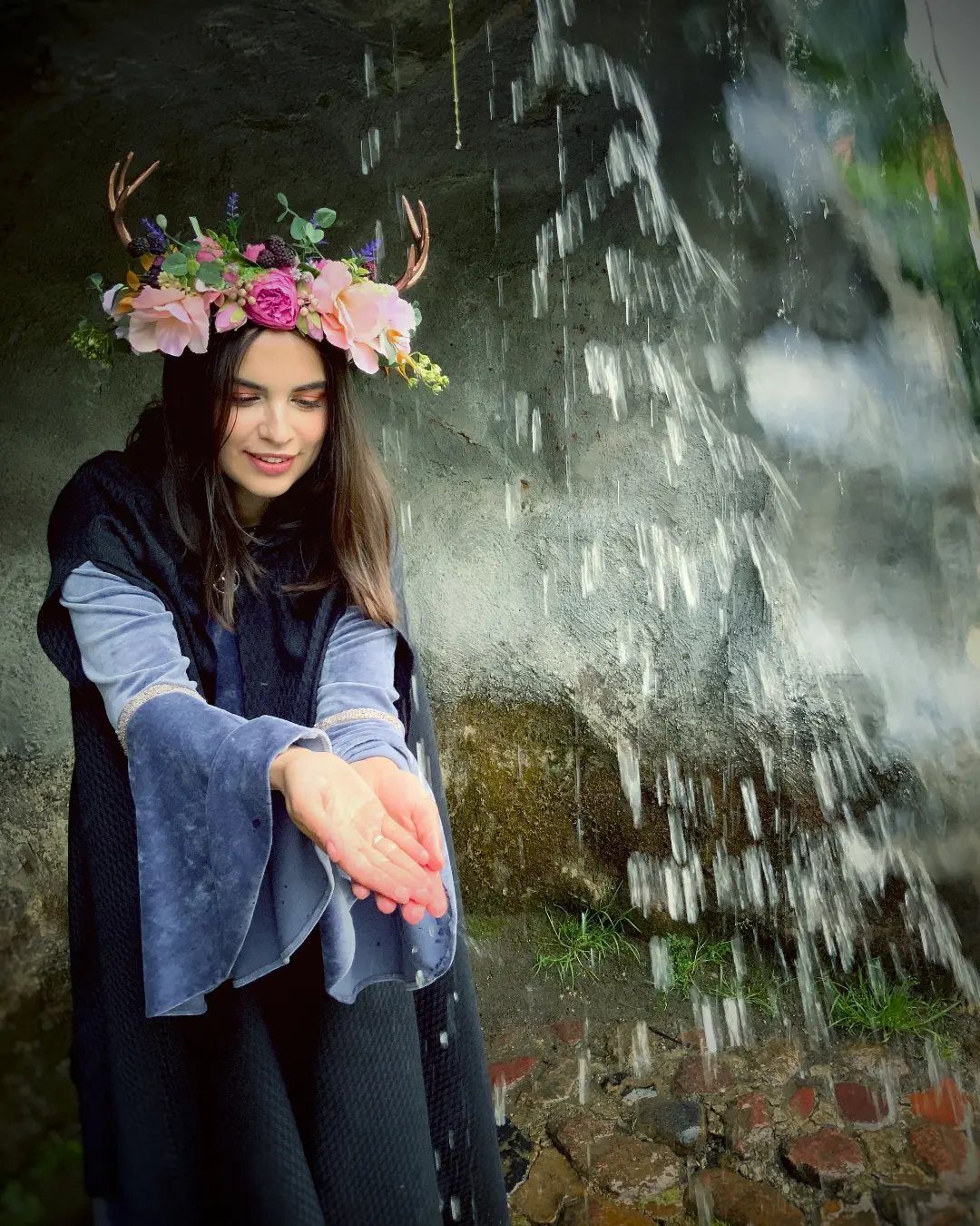 A woman wearing a floral crown with antlers standing by a waterfall.