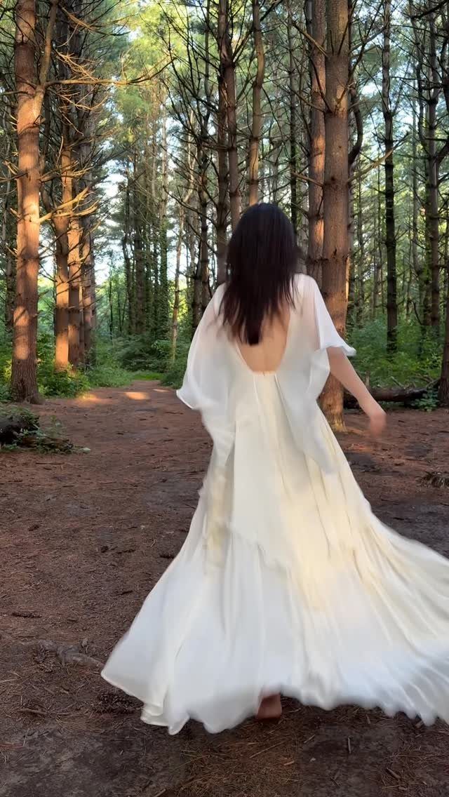 A woman in a flowing white ethereal wedding dress walking through a sunlit forest.