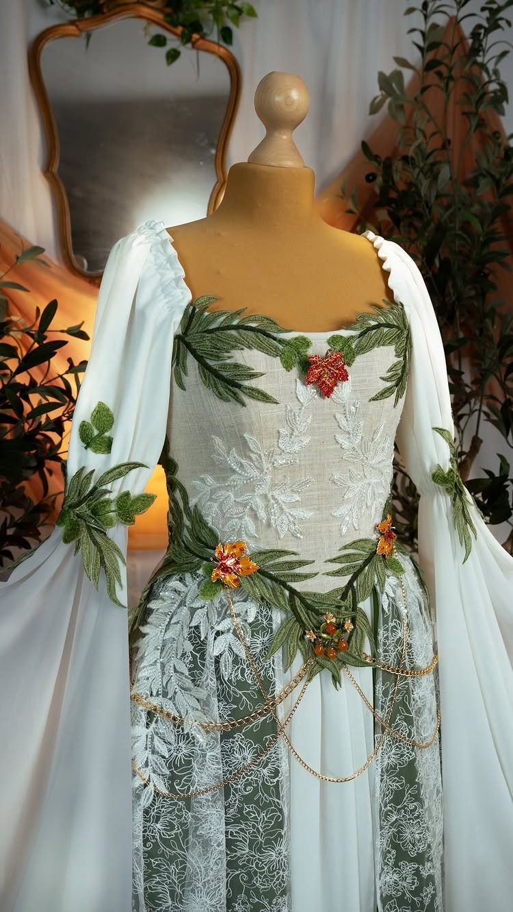 A white fantasy-style wedding dress with green leaf embroidery and gold chains on a mannequin.