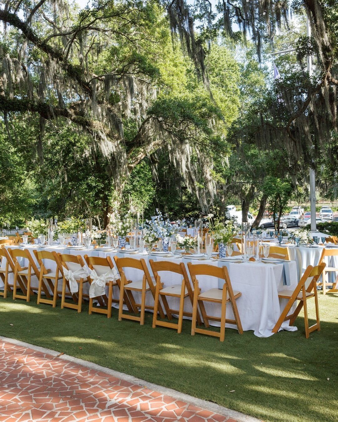 Mansion Wedding Venue Ideas: 17 Estate Settings for 2026 9 Long dining tables with blue runners and floral centerpieces set under mossy trees on a lawn.