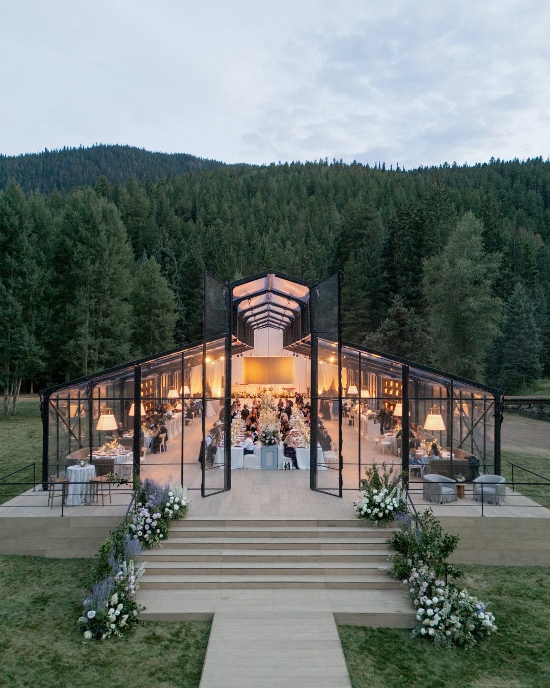 15 Most Beautiful Wedding Venues for 2026 7 A transparent glass wedding marquee tent illuminated at dusk against a backdrop of evergreen forest hills.