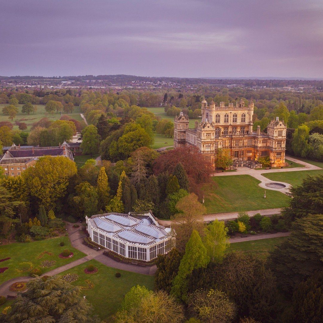 15 Most Beautiful Wedding Venues for 2026 13 A large historic stone mansion and a white glass conservatory surrounded by dense green trees and gardens.