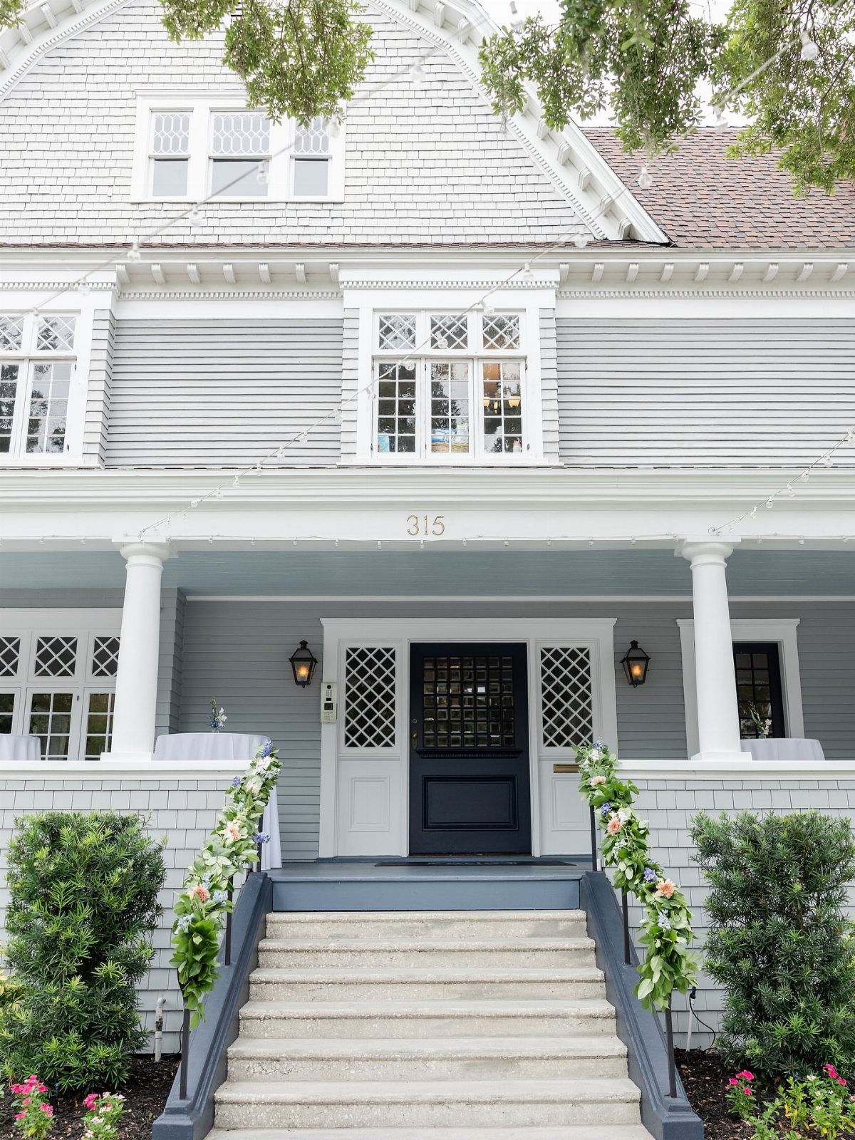 15 Most Beautiful Wedding Venues for 2026 1 A light grey house with white columns and a front porch decorated with floral garlands.