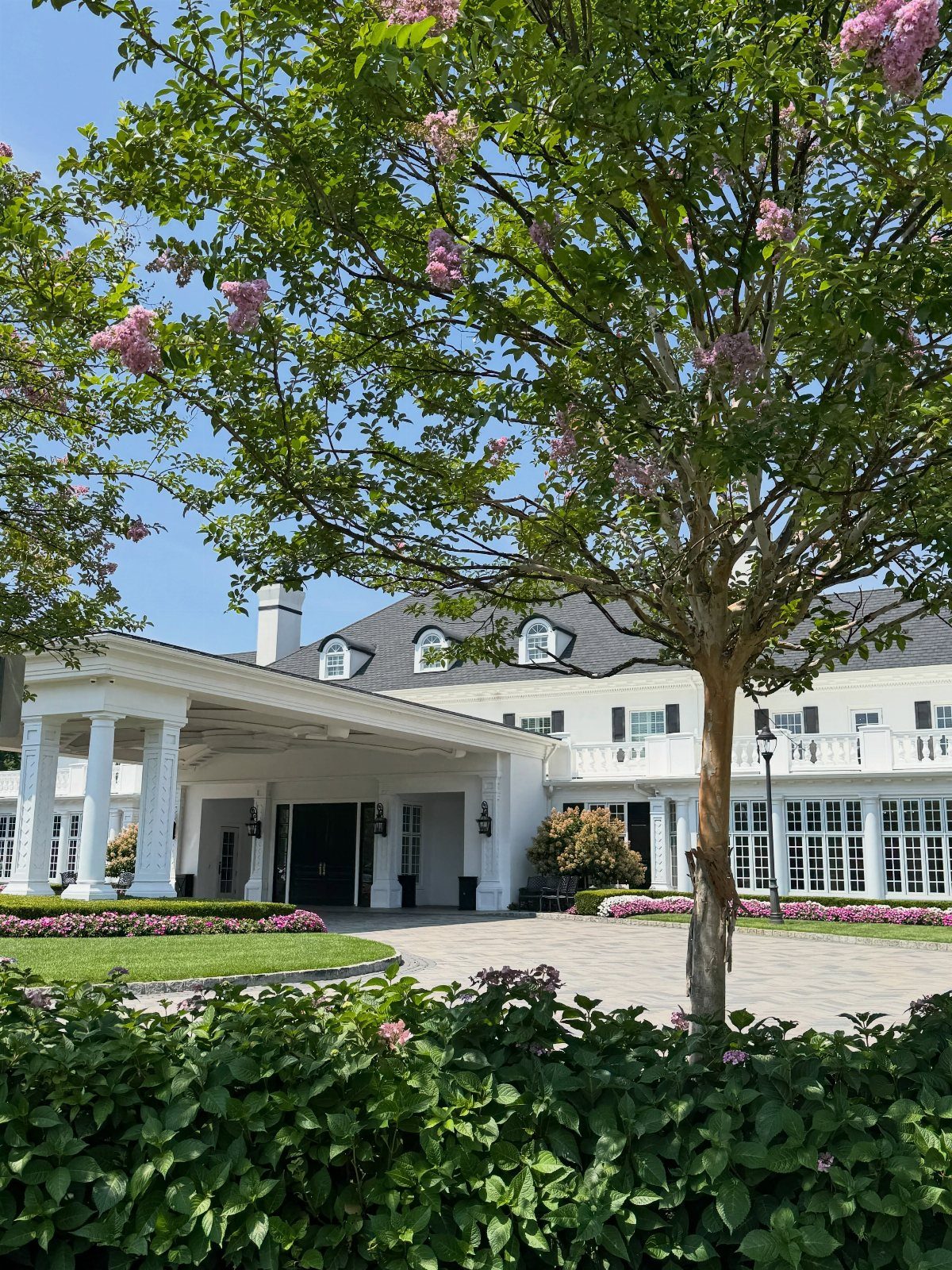 15 Most Beautiful Wedding Venues for 2026 3 A large white estate mansion with columns and flowering trees under a bright blue sky.