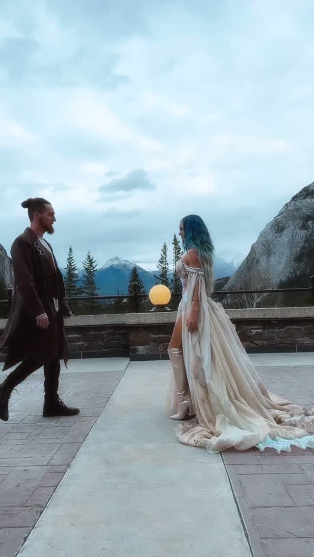 A woman in a flowing cream gown and boots stands on a mountain terrace facing a man.