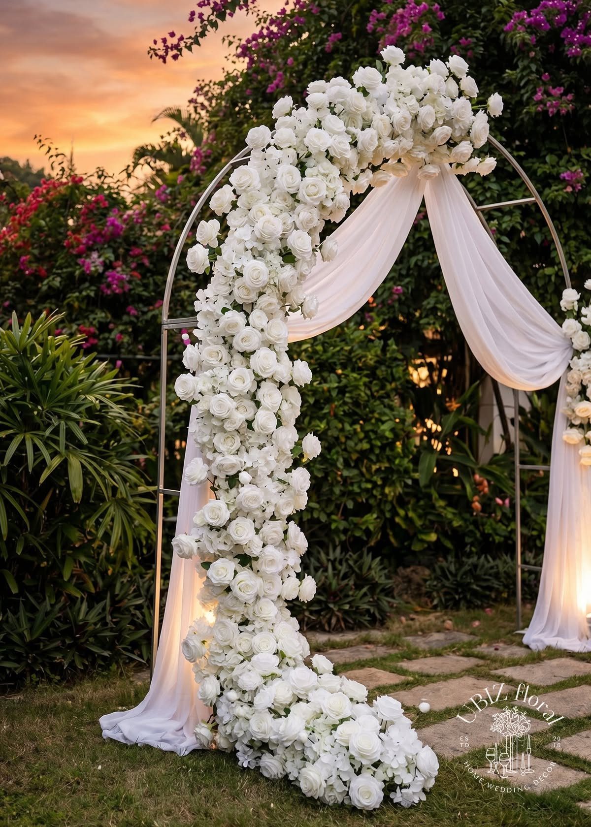 A silver wedding arch decorated with white roses and draped white fabric in a garden.