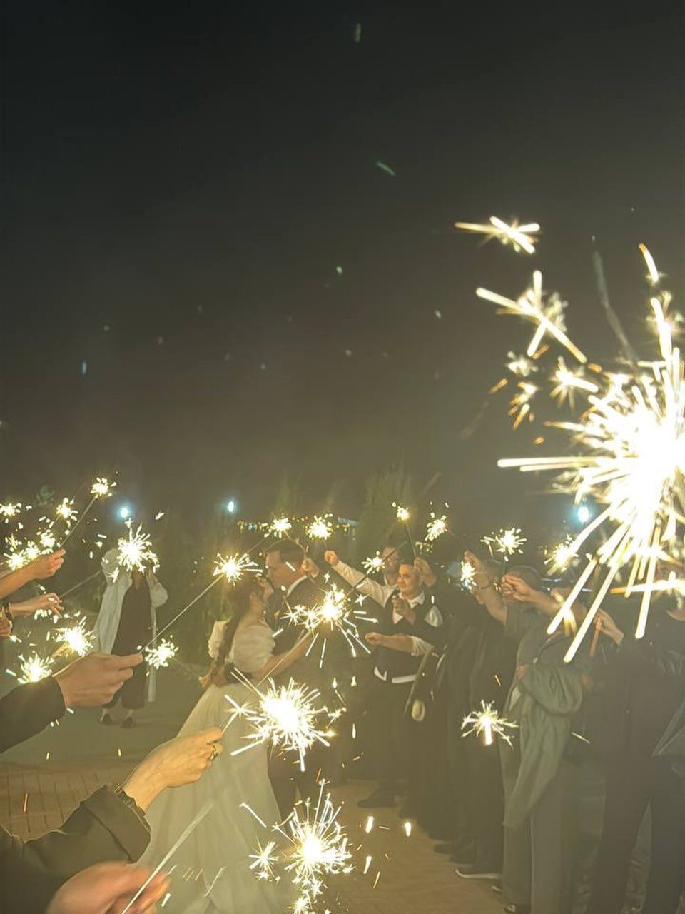A bride and groom walking through a crowd of guests holding bright, glowing sparklers at night.