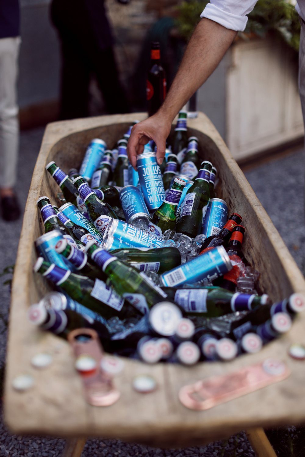 A hand reaching into a wooden trough filled with ice and assorted bottled drinks.