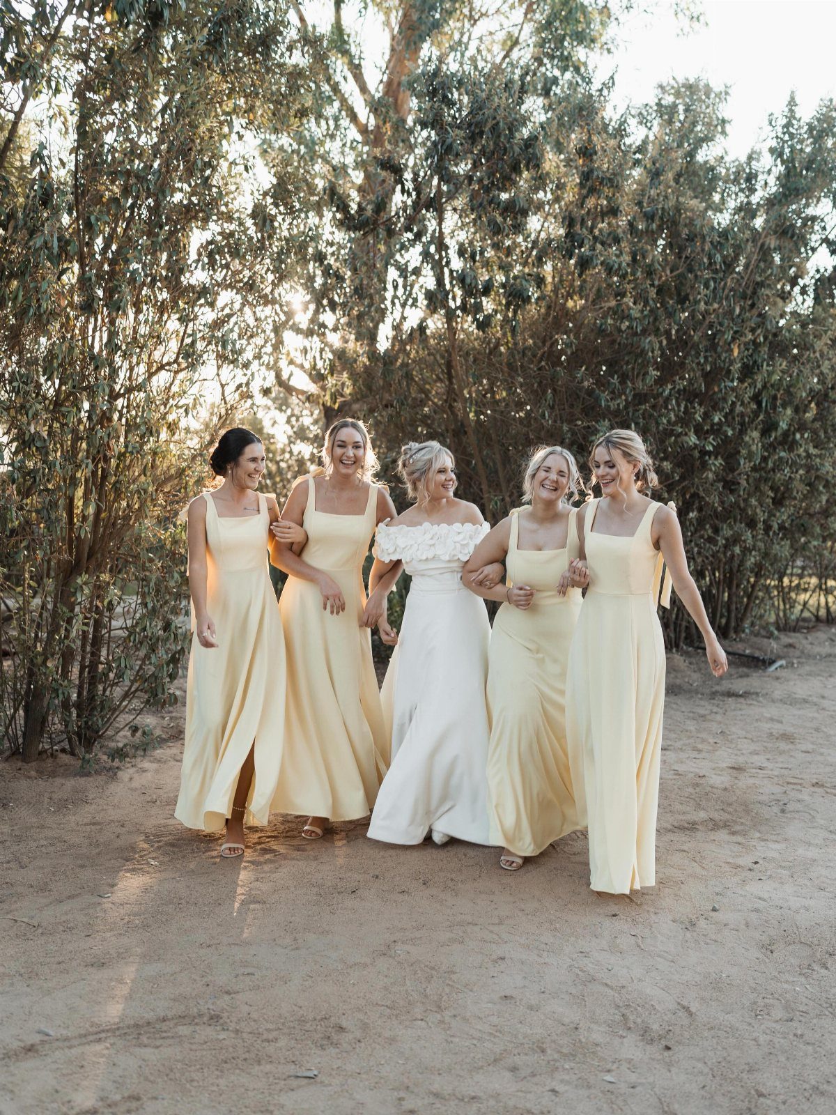 Five women in long dresses walking through a sunlit forest at sunset.