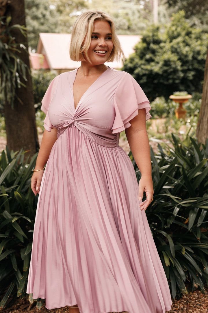A smiling blonde woman wearing a dusty rose pleated midi dress standing in a garden.