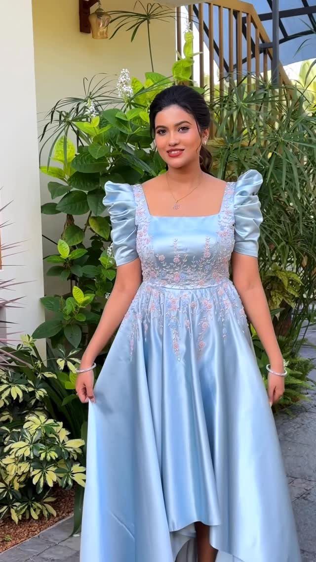 A woman wearing a light blue satin dress with floral embroidery standing among green plants.