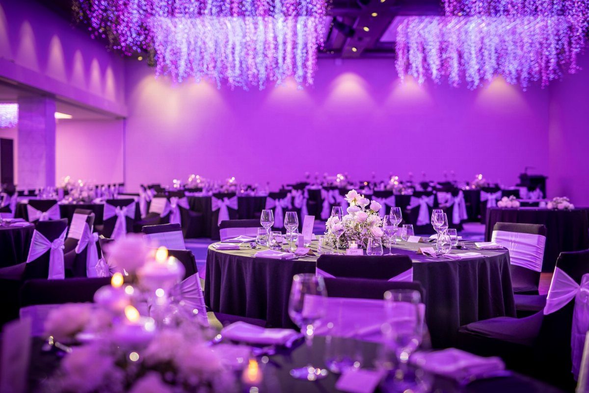 Purple and Black Wedding Theme: 8 Ideas That Actually Work 1 Banquet hall decorated with purple uplighting, black linens, and hanging floral arrangements for a wedding.