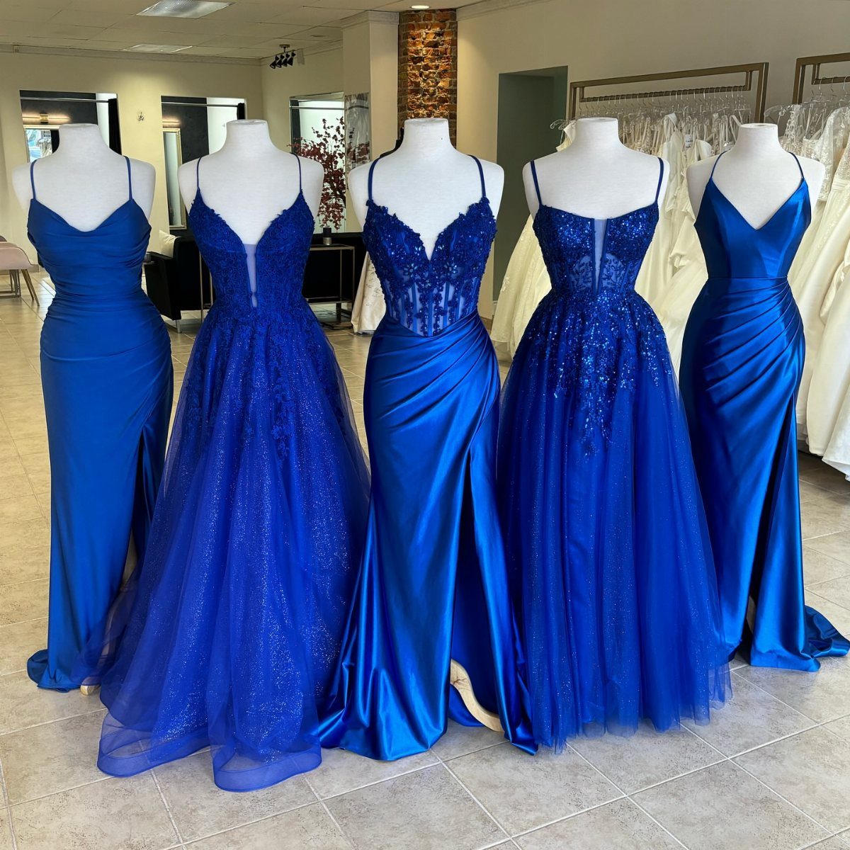 Five different styles of royal blue bridesmaid dresses displayed on white mannequins in a shop.