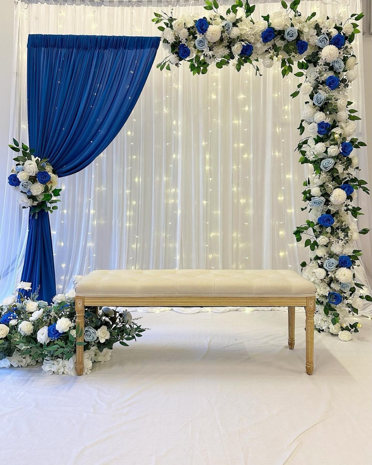 Wedding backdrop with blue drapery, white fairy light curtains, and a floral arch with a cream bench.