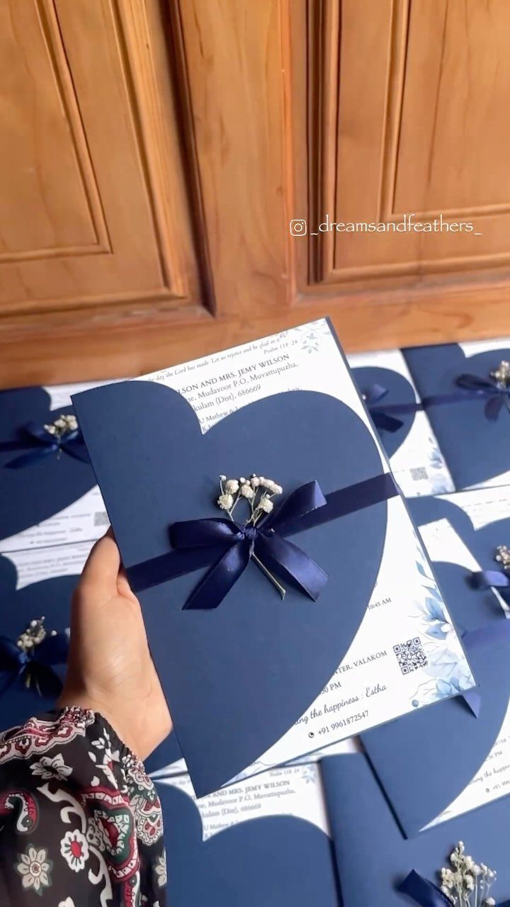 A hand holding a royal blue heart-shaped wedding invitation with a blue ribbon and white flowers.