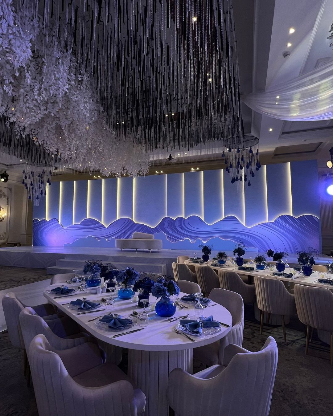 Wedding reception tables set with royal blue napkins and glassware under a silver fringe ceiling installation.