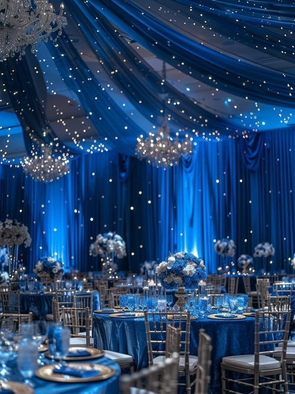 Royal blue draped ceiling with fairy lights and chandeliers over decorated wedding reception tables.