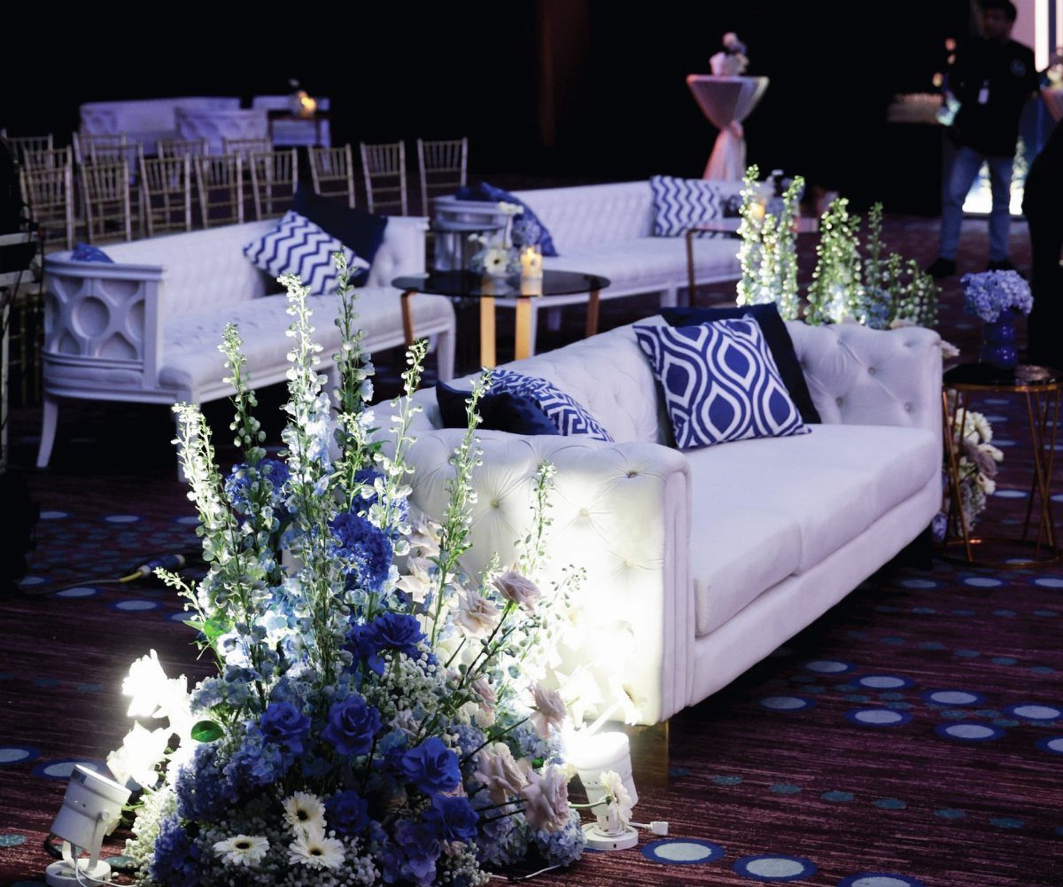 White tufted sofas with royal blue patterned pillows and blue floral arrangements in a wedding lounge.