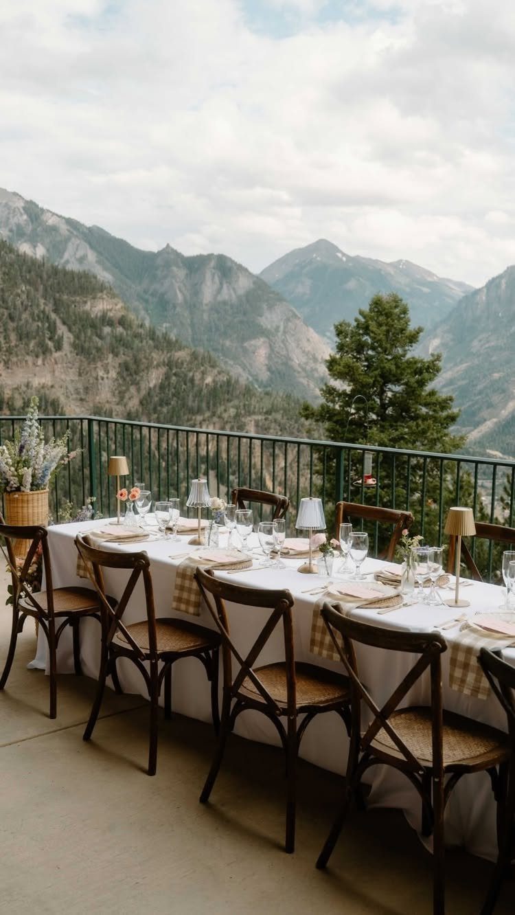 A long dining table set for a meal on a balcony overlooking large mountains.