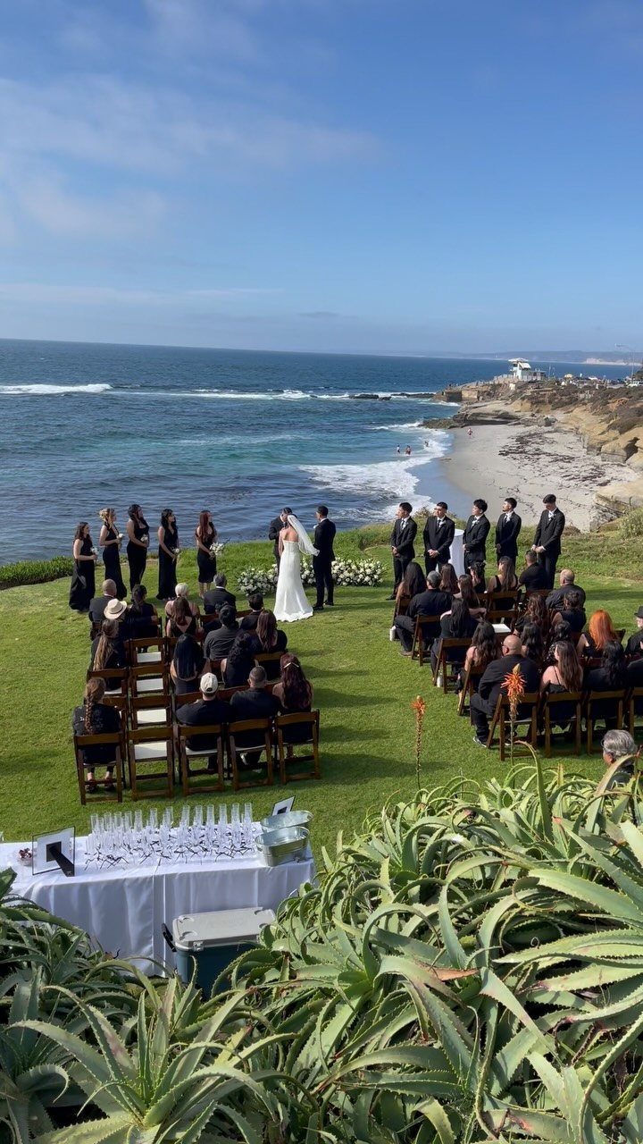 A wedding ceremony on a grassy cliff overlooking the blue ocean waves under a clear sky.
