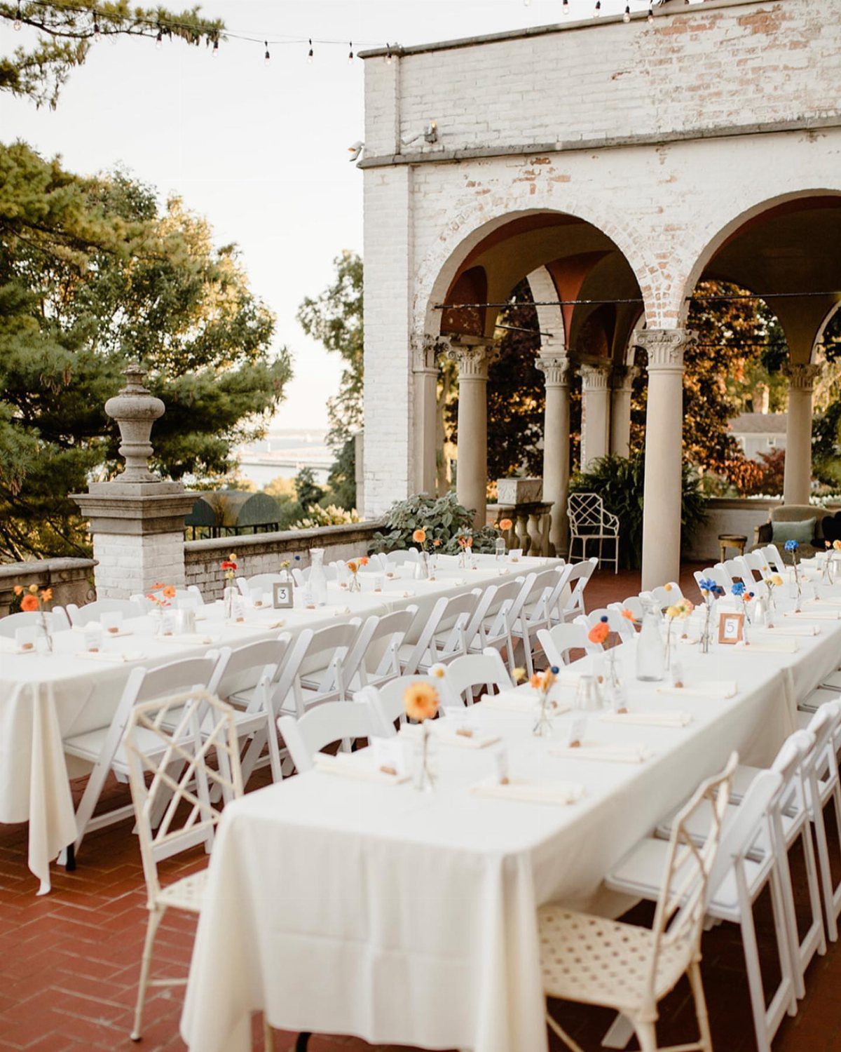Long dining tables with white linens and chairs set on an outdoor patio under arches.
