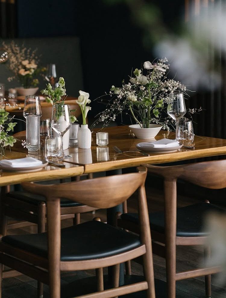 A set wooden dining table with white floral arrangements, glassware, and modern chairs in a restaurant.