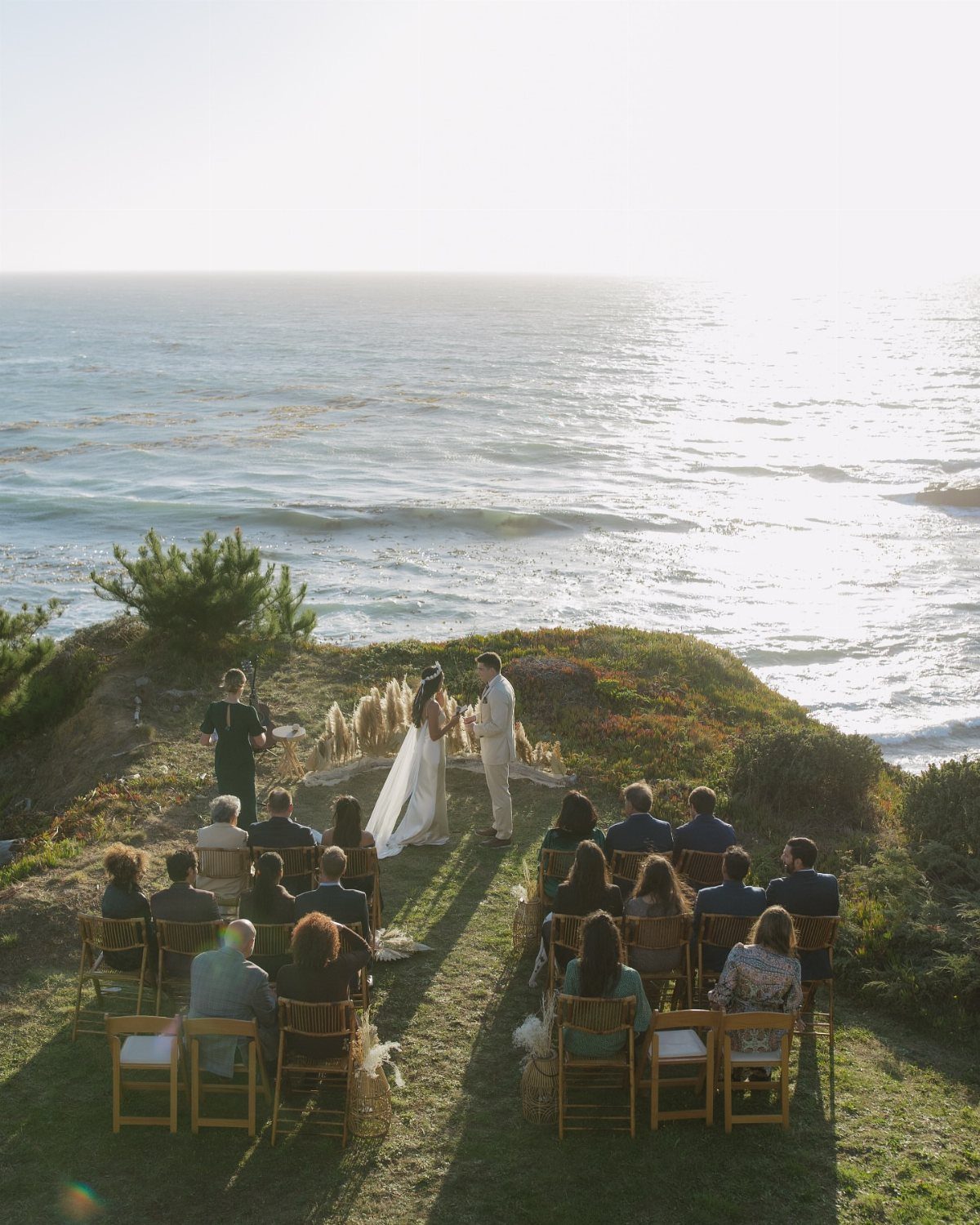 A wedding ceremony on a coastal cliff overlooking the ocean with guests seated in rows.
