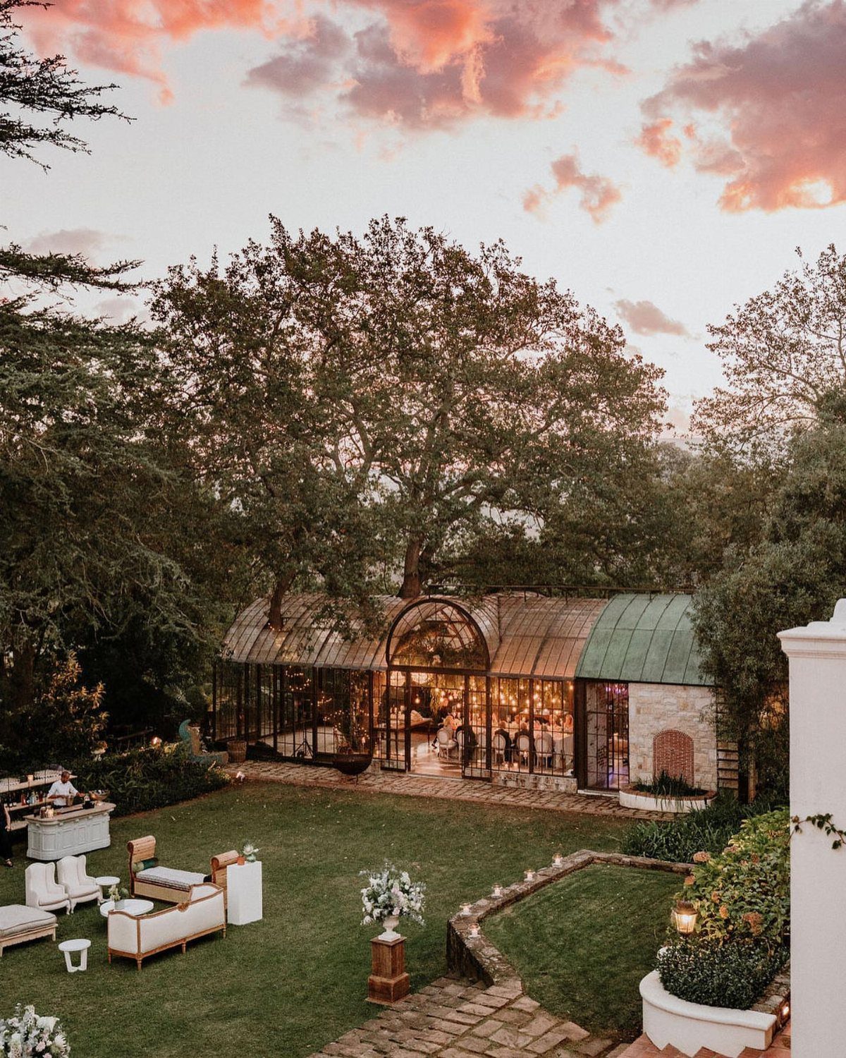 A glass greenhouse wedding venue glowing with warm lights under a sunset sky.