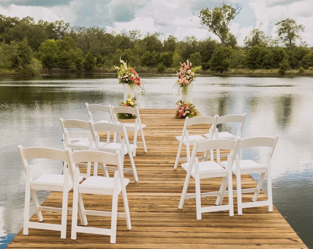 White folding chairs arranged on a wooden dock facing floral arrangements over a calm lake.