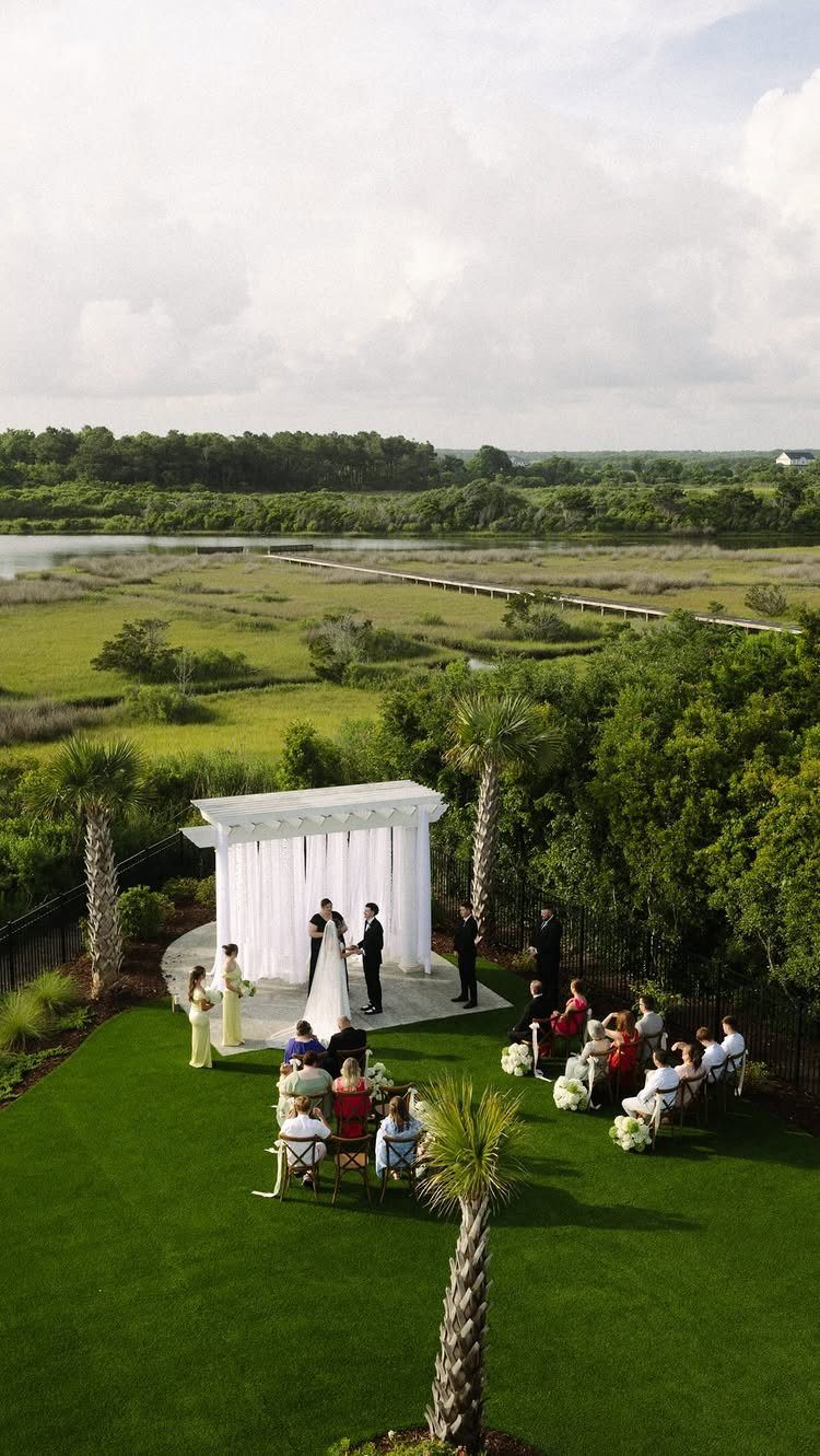 A small wedding ceremony taking place on a green lawn under a white pergola overlooking a marsh.