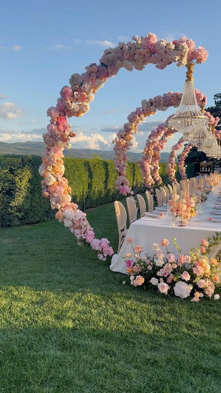 Floral arches frame a long, elegantly set outdoor dining table on a green lawn.