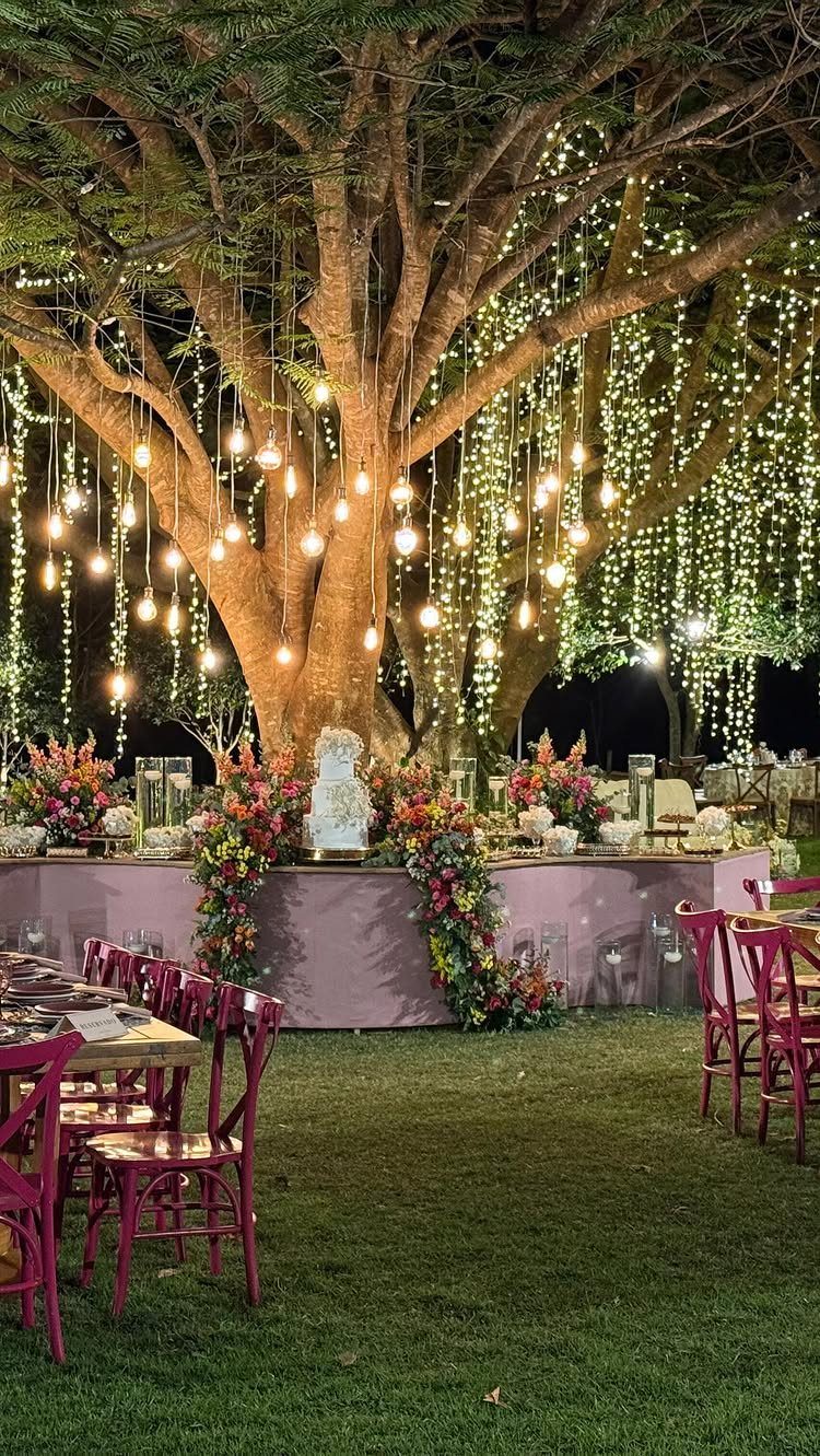 Twilight Forest Wedding Ideas: 10 Concepts for 2026 8 Wedding reception table set under a large tree with hanging lights and colorful floral arrangements.